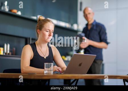 Berlino, Germania. 13 agosto 2024. Foto simbolica sul tema della divisione del lavoro tra coppie in famiglia. Una donna siede in cucina sul suo computer portatile mentre un uomo lava i piatti sullo sfondo. Berlino, 13 agosto 2024. || versione modello credito disponibile: Notizie dpa/Alamy Live Foto Stock