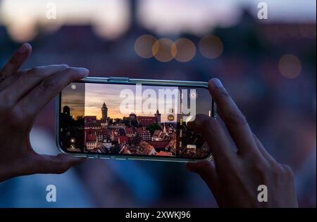 Smartphone Nürnberger Stadtbild am Abend Eine Person fotografiert die Nürnberger Altstadt bei Sonnenuntergang mit einem. Auf dem display ist die Kaiserburg in Nürnberg deutlich zu sehen, während der Hintergrund unscharf bleibt und in bokeh-Lichtern verschwimmt. Die Szene verbindet moderne Technologie mit historischer Architektur. Nürnberg Bayern Deutschland *** paesaggio urbano di Norimberga la sera Una persona fotografa la città vecchia di Norimberga al tramonto con uno smartphone il Castello Imperiale di Norimberga è chiaramente visibile sul display, mentre lo sfondo rimane sfocato e sfocato nel bokeh Foto Stock