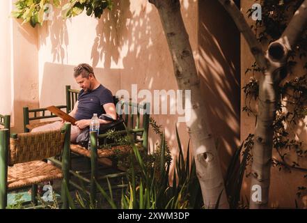 Un adulto medio, abbigliamento casual, uomo caucasico seduto all'aperto all'ombra sotto un albero guardando il menu, in un tranquillo giardino di lusso. Marrakech, Marocco Foto Stock