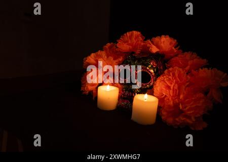 Un cranio nero ornato da fiori metallici, circondato da una vivace corona di calendula arancione. Fiancheggiato da candele accese su sfondo nero, cattura t Foto Stock