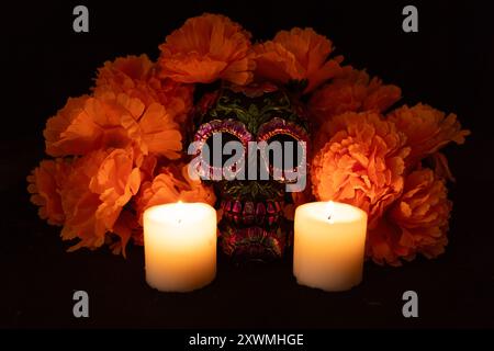 Un cranio nero decorato con fiori metallici, circondato da una vivace corona di calendula arancione. Le candele accese sono posizionate su entrambi i lati, appoggiate a una profondità di Foto Stock