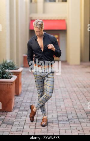 Un bel biondo con una camicia nera e pantaloni a quadri cammina con sicurezza su una strada di mattoni Foto Stock