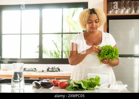 Giovane donna afroamericana curvy in piedi in cucina a preparare un'insalata Foto Stock