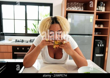 Giovane donna afroamericana curvy in cucina che si diverte a mangiare un hamburger grasso Foto Stock