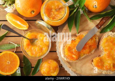 Marmellata d'arancia fatta in casa su fette di pane e vasetti pieni di marmellata e frutta intorno al tavolo di legno. Vista dall'alto. Foto Stock