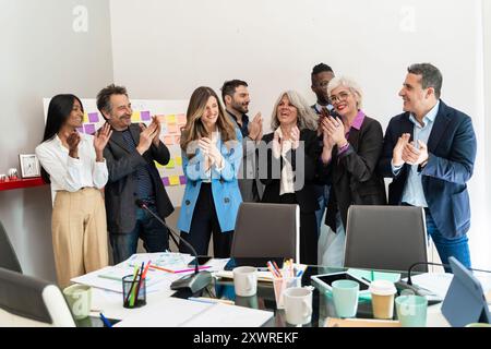 Team aziendale entusiasta e diversificato che plaude a una presentazione di successo. Professionisti multigenerazionali in un ufficio moderno con appunti e disordine Foto Stock