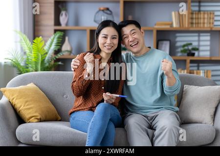 Gioiosa coppia asiatica che celebra il risultato positivo del test di gravidanza sul divano. Felicissima coppia matura che esprime entusiasmo e felicità nell'aspettarsi il bambino, creando un'atmosfera gioiosa e di speranza Foto Stock
