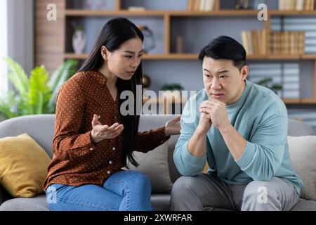 Coppia asiatica in discussione emotiva sul divano. La donna gesta con preoccupazione mentre l'uomo ascolta con attenzione, riflettendo una comunicazione ponderata e un legame familiare. La scena trasmette emozioni relazione Foto Stock