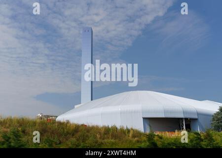 Alto camino bianco sopra un edificio industriale con tetto curvo, adagiato contro il cielo blu e le nuvole bianche. Fabbrica di knoll erboso con alberi davanti Foto Stock