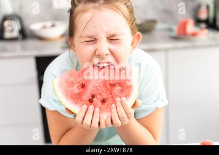 Una bambina morde un pezzo di anguria mentre si trova in cucina a casa con una morbida t-shirt turchese. Foto di alta qualità Foto Stock