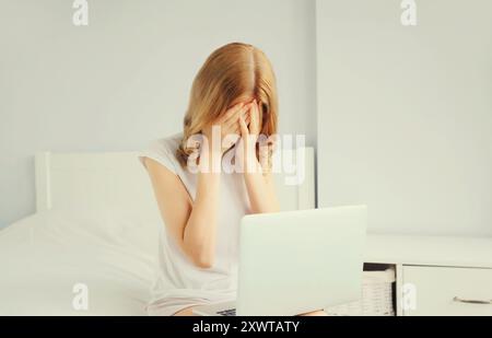 Ritratto di una giovane donna triste e turbata che lavora con un computer portatile sul letto in una stanza bianca di casa Foto Stock