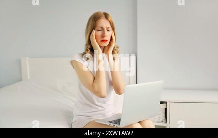 Ritratto di una giovane donna stanca con mal di testa lavorava con un portatile sul letto in una stanza bianca a casa Foto Stock