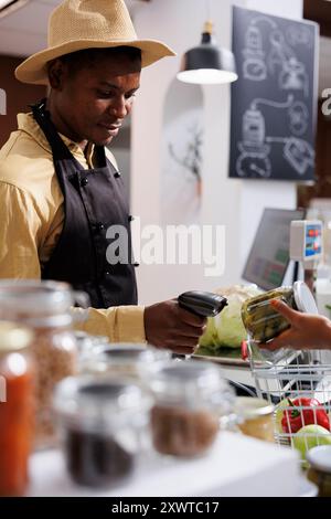 Il negoziante afroamericano maschile al supermercato ecologico promuove lo shopping sostenibile e offre prodotti freschi, biologici e privi di plastica. Immagine dettagliata di un cassiere che scansiona gli articoli al bancone. Foto Stock