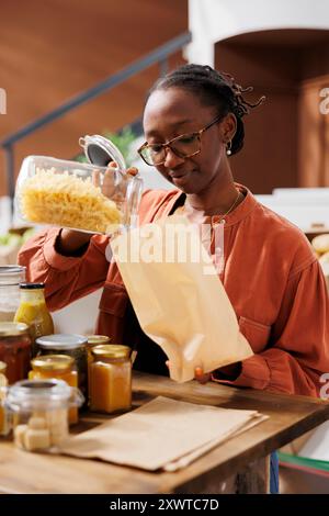 Il ritratto di donna nera con gli occhiali sta versando pasta fusilli in un sacchetto di carta marrone. Giovani spaghetti di farcitura afro-americana appena fatti in un sacco senza plastica in un minimarket ecologico. Foto Stock