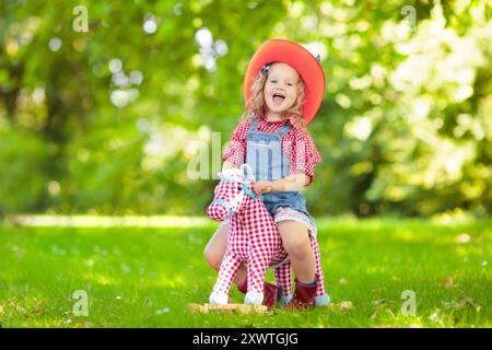 Bambino e bambina vestiti da cowboy e cowgirl che giocano con il cavallo a dondolo giocattolo nel parco. I bambini giocano all'aperto. Bambini in costumi di Halloween Foto Stock