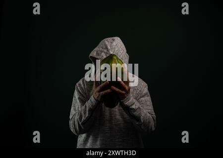 Uomo misterioso con occhiali con cappuccio che tiene la papaya verde davanti al viso. Isolato su sfondo scuro. Foto Stock