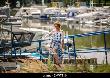 Una donna passeggia lungo una ringhiera blu vicino a un porticciolo pieno di barche, godendosi la calda luce del sole e gli splendidi dintorni. Foto Stock