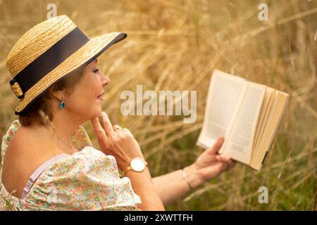 Una donna con un cappello di paglia ama leggere in un campo dorato illuminato dal sole circondato da erba alta. Foto Stock