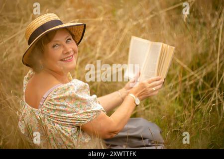 Una donna con un cappello di paglia sorride mentre legge un libro in un campo illuminato dal sole, circondato da erba dorata in una calda giornata estiva. Foto Stock
