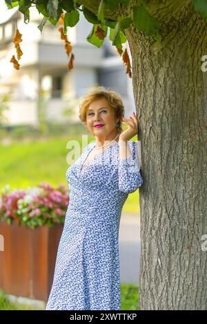 Una donna con i capelli ricci gode un momento sereno accanto a un albero, indossando un abito azzurro e sorridendo tra fiori vivaci. Foto Stock