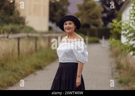 Un'elegante donna passeggia lungo un percorso in giardino, vestita con una blusa bianca a spalla e una gonna nera fluente, con il verde che la circonda. Foto Stock