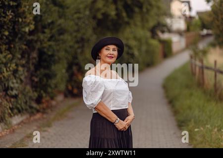 Una donna che indossa un'elegante blusa bianca e una gonna nera si erge in tutta sicurezza lungo un percorso alberato in una giornata di sole. Foto Stock
