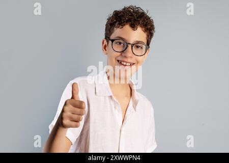 Ritratto di un giovane allegro con capelli ricci che indossano occhiali e camicia bianca, che mostra un gesto con i pollici in alto. Concetto di positività, approvazione e confid Foto Stock