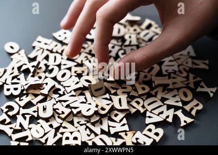 Primo piano di dita che raccolgono lettere dell'alfabeto di legno sparse su una superficie scura. Concetto di comunicazione, istruzione, giochi di parole e apprendimento delle lingue. Foto Stock