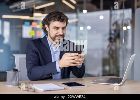 Uomo d'affari sorridente mentre utilizza uno smartphone in un ufficio moderno, circondato da un notebook e un tablet, che mette in mostra le comunicazioni digitali e il multitasking. Foto Stock