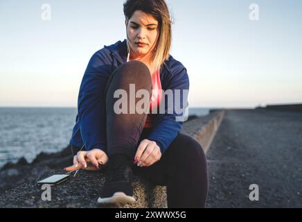 Una giovane donna in sovrappeso si sta preparando ad allenarsi sul molo mentre si legano le scarpe da ginnastica. Ha un cellulare e indossa gli auricolari. Foto Stock