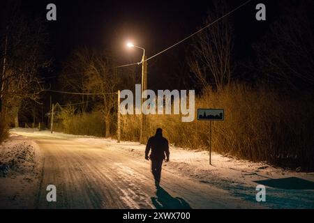 Una figura solitaria cammina lungo una strada innevata, illuminata da un unico semaforo nella quiete della notte. Questa immagine cattura l'essenza della solitudine e dell'introspezione, ricordando i tranquilli viaggi attraverso la foresta di Białowieża. Foto Stock