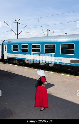 Una donna in un vivace abito rosso tradizionale ungherese si trova in una stazione ferroviaria. Il suo abbigliamento elegante contrasta con il moderno treno blu dietro di lei, catturando un mix di cultura e viaggi. La scena è ambientata sotto un cielo azzurro, sottolineando il fascino senza tempo della moda tradizionale ungherese. Foto Stock