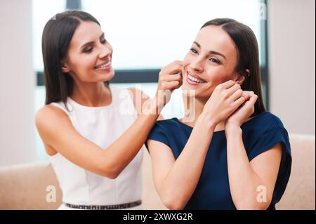 Preparatevi a uscire di notte. Bella giovane donna in abito da sera che aiuta la sua amica con gli orecchini e sorride mentre entrambi sono seduti sul divano Foto Stock