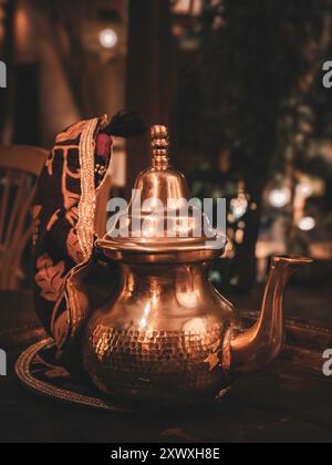 Una teiera in metallo marocchino splendidamente lavorata con disegni intricati, appoggiata su un rustico tavolo in legno. Lo sfondo è leggermente illuminato, creando un ambiente caldo Foto Stock