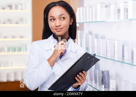 Sto pensando a una medicina adeguata per te. Giovane donna africana premurosa con un camice da laboratorio che tiene gli appunti e guarda con sorriso mentre è in piedi vicino Foto Stock
