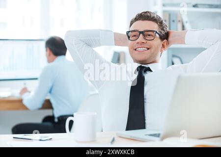 Soddisfatto del lavoro svolto. Allegro uomo d'affari in abbigliamento formale che tiene la testa in mano mentre siede alla sua scrivania in ufficio Foto Stock