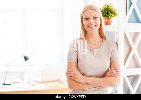 Allegra donna d'affari. Bella giovane donna appoggiata allo scaffale e sorridente mentre si trova vicino al suo posto di lavoro in ufficio Foto Stock