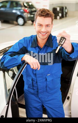 Prenda la sua macchina. Giovane allegro in uniforme che si allunga mano con le chiavi mentre si trova vicino alla vettura in officina Foto Stock