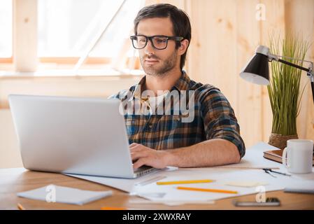 Tecnologia moderna per il lavoro creativo. Giovane sicuro di sé che lavora su un notebook mentre è seduto sul posto di lavoro Foto Stock