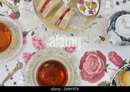 Festa retrò del tè britannico alle cinque: Due tazze di teiera, un cupcake con panna e frutta su un altopiano, panini triangolari su un tavolo. Vista dall'alto. Foto Stock