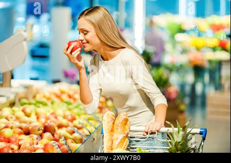 Solo la migliore frutta e verdura. Bella giovane donna che tiene in mano la mela e la annusa con sorriso mentre si trova in un negozio di alimentari Foto Stock