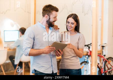 Guarda questo! Giovane uomo e donna allegri che discutono di qualcosa e guardano insieme un tablet digitale mentre i loro colleghi lavorano dietro Foto Stock