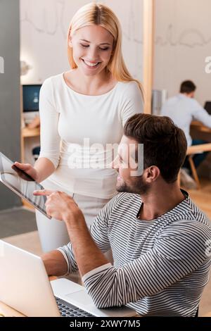 Chiedere un Consiglio di un esperto. Giovane uomo e donna allegri che discutono di qualcosa e guardano insieme un tablet digitale mentre i loro colleghi lavorano con me Foto Stock