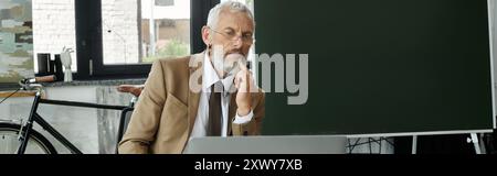 Un uomo maturo con la barba seduto davanti a un laptop, insegnando con attenzione online, insegnante lgbtq Foto Stock