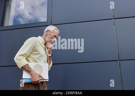 Un uomo di mezza età con la barba tiene i documenti e guarda con attenzione al terreno di fronte a un edificio moderno. Foto Stock