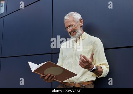 Un uomo barbuto con una camicia gialla sorride mentre legge un libro di fronte a un edificio moderno. Foto Stock