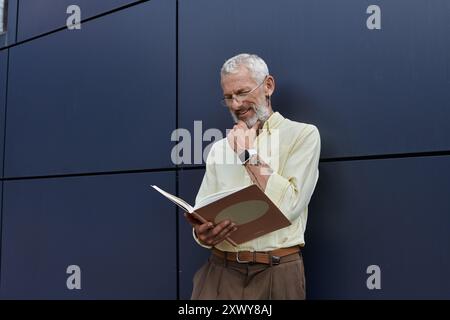 Un gay maturo con la barba legge un libro mentre si trova accanto a un edificio moderno, toccando con cura il mento. Foto Stock