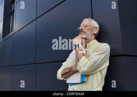 Un uomo gay maturo con barba e occhiali si trova vicino a un edificio moderno, che tiene una pila di libri. Foto Stock