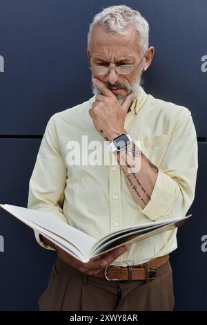 Un uomo maturo con barba e occhiali legge attentamente un libro mentre si trova vicino a un edificio moderno. Foto Stock