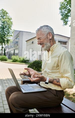 Un uomo maturo con la barba seduto su una panchina, sorridendo mentre usa il suo portatile. Indossa una camicia giallo chiaro e ha un tatuaggio sul braccio. Foto Stock